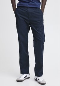 Tumši zilas, vieglas taisnas bikses ar elastīgu jostu un sānu kabatām, apvienotas ar pelēkiem un baltajiem sneaker apaviem ar melnām svītrām.