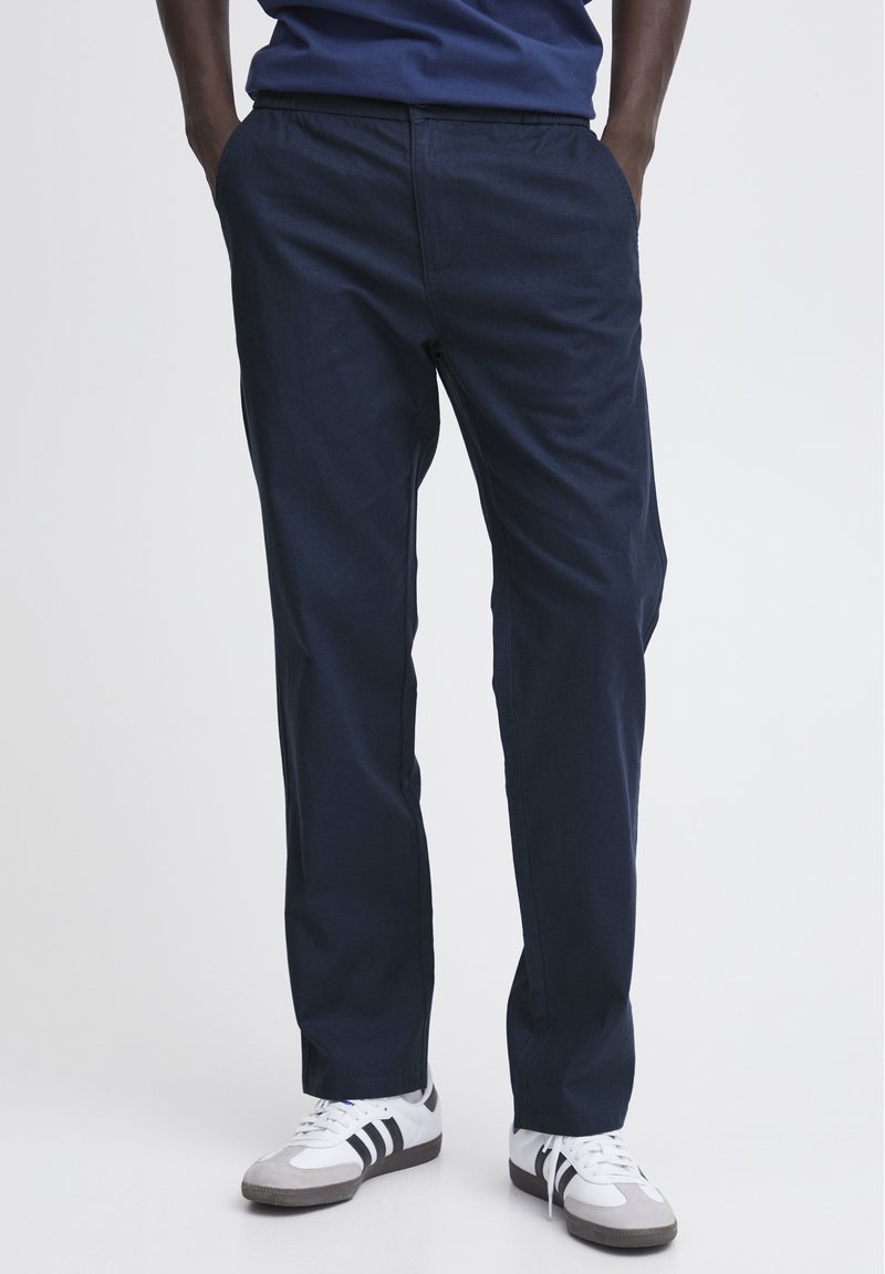 Tumši zilas, vieglas taisnas bikses ar elastīgu jostu un sānu kabatām, apvienotas ar pelēkiem un baltajiem sneaker apaviem ar melnām svītrām.