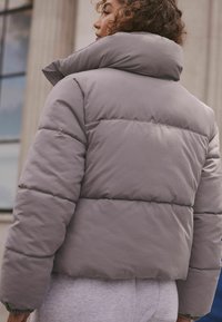 Pelēks pūkains jaka ar augstu apkakli, uzšūtu struktūru un īsu dizainu. Redzamā šūšana un mīksta auduma nodrošina siltumizolāciju.