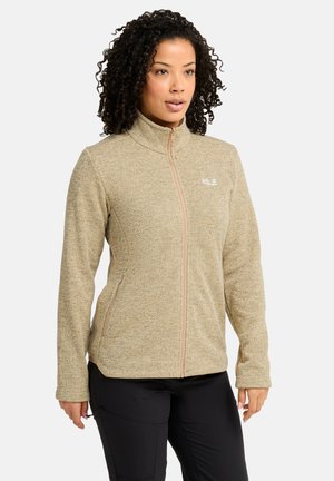 Frau mit lockigem Haar, die eine beige Fleecejacke mit Reißverschluss und schwarze Hose trägt, schaut leicht zur Seite mit neutralem Gesichtsausdruck.
