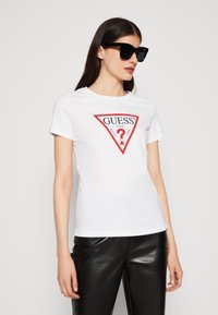 Maglietta bianca con logo a triangolo rosso che presenta il testo "GUESS?". Maniche corte, colletto rotondo, abbinata a pantaloni neri lucidi.