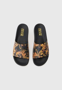 Sandálias de slide pretas com um upper brilhante e um padrão dourado ornamental. Texto "VERSACE JEANS COUTURE" em amarelo na palmilha.