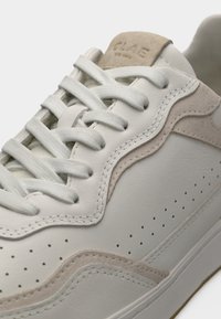 Κοντινή λήψη μπεζ και υποκίτρινου αθλητικού παπουτσιού με υφασμάτινα κορδόνια, διατρητικές πλευρές και σουέντ λεπτομέρειες γύρω από τις άκρες και τη γλώσσα.