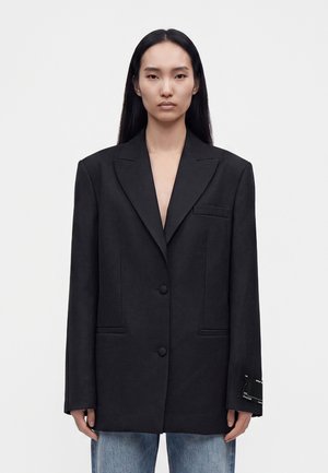 Vrouw met lang zwart haar draagt een oversized zwarte blazer en blauwe jeans, staand tegen een eenvoudige witte achtergrond.