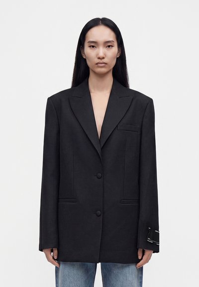 Femme aux longs cheveux noirs porte un blazer noir oversize et un jean bleu, debout devant un fond blanc uni.