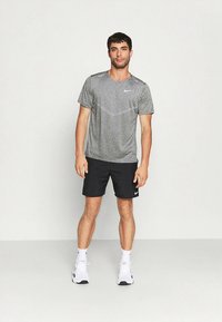 Moška športna obleka, ki vključuje sivo, kratkoročno teksturirano majico in črne kratke hlače, združene z belimi supergami.