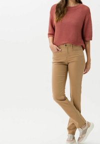 Mujer vistiendo un suéter de punto color óxido, pantalones beige y zapatillas blancas beiges, de pie con una mano en el bolsillo frente a un fondo plano.