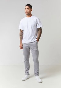 Mann med tatoverte armer iført en hvit T-skjorte, lysegrå bukser og hvite sneakers som står mot en ensfarget lys bakgrunn.