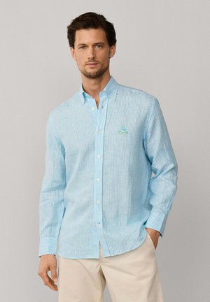 Homme portant une chemise boutonnée à rayures bleu clair et blanc avec un pantalon beige, debout devant un fond gris uni.