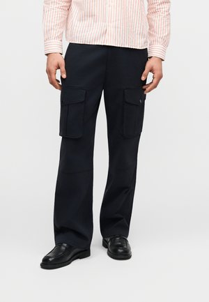 Άνδρας που φοράει μαύρο παντελόνι cargo με πλαϊνές τσέπες με καπάκι, μαύρα παπούτσια και λευκό πουκάμισο μακρυμάνικο με ροζ ρίγες, σε απλό φόντο.