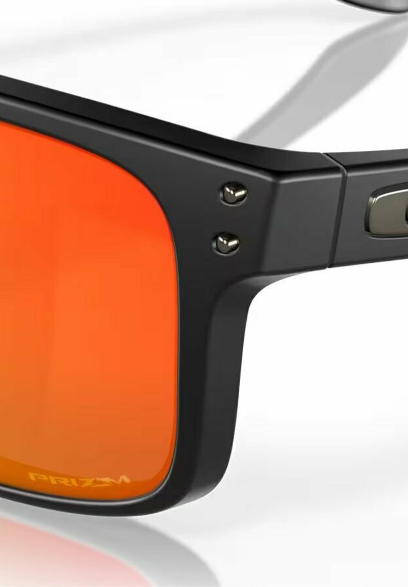 Lunettes de soleil noires avec des verres orange et une finition mate. Détails remarquables : accents argentés sur la monture et un logo sur le verre.