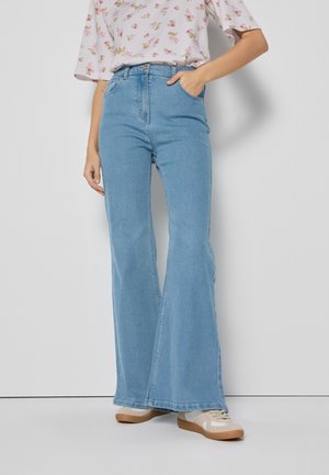 Jasnoniebieskie dżinsy w kształcie dzwonów z wysokim stanem, wykonane z denimu. Tkanina ma subtelną teksturę. Noszone z beżowymi trampkami i kwiecistą bluzką.