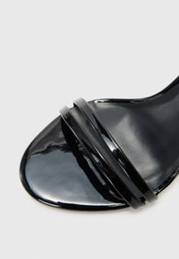 Schwarze Lackledersandale mit einer glatten, glänzenden Oberfläche, die zwei schwarze Akzentriemen über dem Zehenbereich und eine konturierte Sohle aufweist.