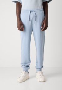 Lys blå joggebukser laget av mykt stoff, med elastisk liv med snøring og tettsittende mansjetter. Brukt med hvite joggesko.
