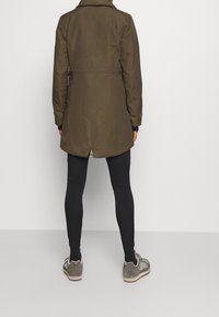 Olivgrön kappa med bälte i midjan, långa ärmar och vent bak, ihop parat med svarta leggings och grå sneakers. Slät textur.