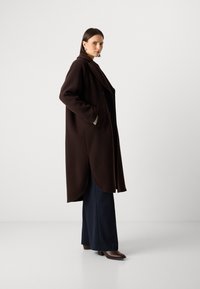 Bruine, oversized jas met een sjaalkraag en zijzakken, gedragen over gestreepte broeken en bruine haklaarzen.