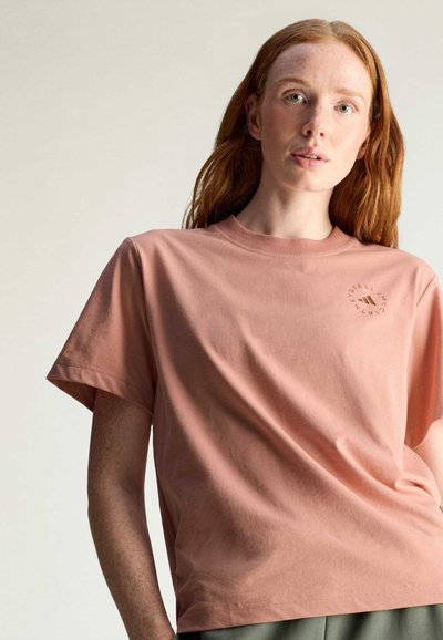 T-shirt in cotone rosa chiaro con maniche corte e scollo rotondo, caratterizzato da una piccola grafica circolare nell'angolo in alto a sinistra.