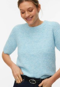 Ženska se smeji, oblečena v svetlomodro teksturirano pulover s kratkimi rokavi, temne kavbojke s črnim pasom, roke počivajo na bokih.