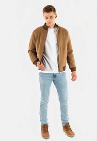 Brun bomberjacka med ribbad krage, kombinerad med en vit t-shirt och ljusblå jeans. Skorna är bruna med snören. Enkel design.