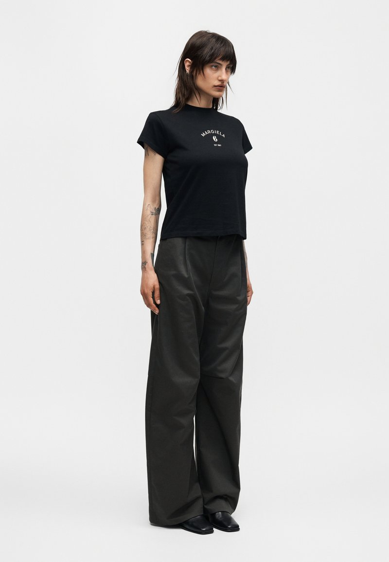 Woman with shoulder-length dark hair wearing black Margiela logo t-shirt, wide-leg black pants, and black shoes against white background.