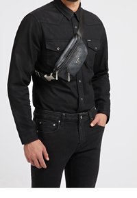Borsa a tracolla in tessuto nero con scomparti con zip, superficie testurizzata e logo triangolare, indossata a tracolla da una persona in camicia nera a pulsante.