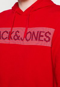 Sudadera roja con cordón, que presenta una franja horizontal blanca y azul marino. El texto "ACK & JONES" está prominentemente exhibido en azul marino sobre la franja.