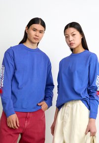 Blaue Langarmshirts mit Rundhalsausschnitt, die grafischen Text auf den Ärmeln zeigen. Ein Modell trägt rote Hosen; das andere Modell trägt cremefarbene Hosen.