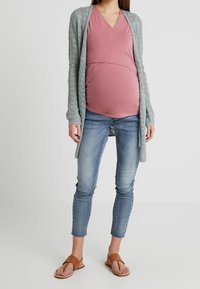 Tenue de maternité comprenant un haut rose en V, un cardigan à motifs bleu clair, un jean ajusté et des sandales marron. Le cardigan a un design ouvert.