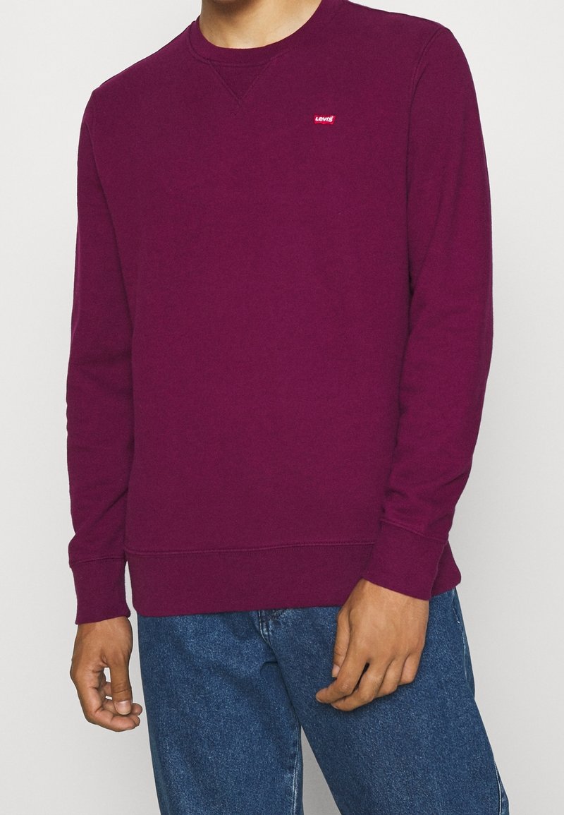 Homme portant un sweat Levi's uni violet foncé à manches longues et un jean bleu, debout, les bras détendus le long du corps.