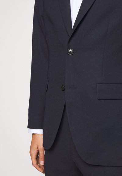 Blazer azul-marinho feito de tecido texturizado, apresentando dois botões, lapela de corte angulado e bolsos laterais. As mangas mostram uma camisa branca por baixo.