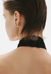 Pendiente de oreja de oro con un diseño ondulado, colocado frente a un peinado corto, acompañado de un choker de tela negra alrededor del cuello, mostrando una piel suave.
