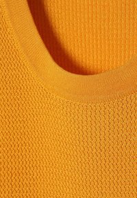 Nærbillede af orange struktureret strikket stof med en rund halsudskæring, som viser detaljeret syning og vævemønstre.