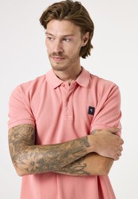 Kurzärmliges, hellrosa Poloshirt aus Baumwolle mit zwei Knöpfen am Ausschnitt und einem kleinen dunklen Patch-Logo auf der linken Brust.