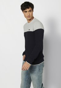 Adīts džemperis gaiši pelēkā, jūras zilā un baltā krāsā. Iespējas audumam ar tekstūru, apaļa kakla izgriezums un ribotas piedurknes ar diskretu logotipa akcentu.