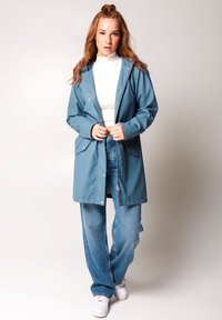 Vedennepropustný modrý plášť do deště so kapucňou, zipsom a dvoma prednými vreckami. V kombinácii s svetlomodrými širokými džínsami a bielymi topánkami.