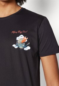 Black t-shirt with small graphic of a flying bee with orange wings, clouds, and text "Flying Buzz-Zaw! Alpha Industries" on the chest.