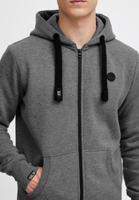Grå zip-up hoodie lavet af blødt materiale, med sorte snortræk, en frontlomme og et rundt logo-detalje på brystet.