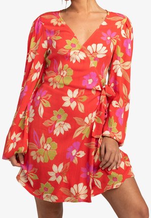 Femme portant une robe portefeuille rouge à manches longues avec un imprimé floral en blanc, rose, vert et violet, debout avec une main posée sur sa cuisse.