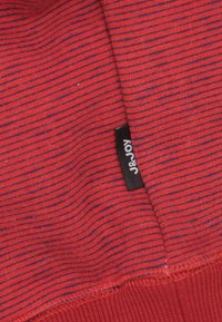 Rood gestreepte stof met blauwe accenten, voorzien van een ribbelstructuur en een zwart etiket met de tekst "J&JOY" nabij de zoom.