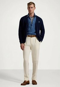 Man som bär en marinblå cardigan, en blå denimskjorta över en randig t-shirt, beige byxor, brun bälte och bruna läderloafers, stående i en neutral miljö.