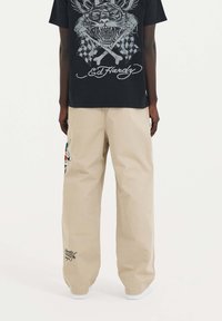 Beżowe spodnie szerokie o gładkiej fakturze, z kolorowym haftem w kształcie węża i detalem logo blisko dołu.