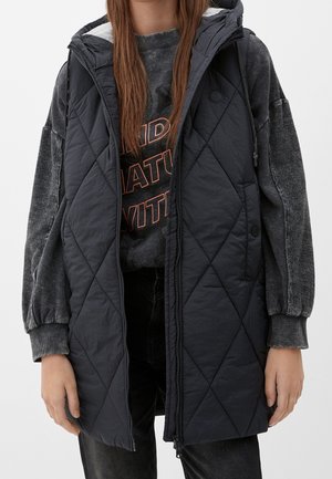 Une personne porte une veste sans manches matelassée noire par-dessus un sweat-shirt gris foncé avec un texte orange, associée à un pantalon noir, debout, les bras détendus.