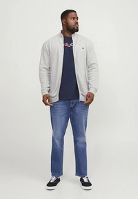 Gaiši pelēks džemperis ar rāvējslēdzēju virs tumši zilas T-krekla, kopā ar zilām džinsa biksēm un melnām sporta kurpēm ar baltiem šņorēm. Ikdienas stils, vienkāršs dizains.