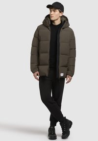 Dark green puffy jacket with a hood, zip closure, and front pocket. Worn over a black sweater and black pants, paired with black sneakers.