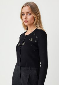Cardigan noir à côtes avec manches longues, arborant des insectes brodés et des motifs floraux. Coupe ajustée et fermeture à boutons sur le devant.