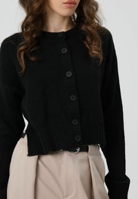 Zwarte gebreide cardigan met een ronde hals en zes knopen sluiting. Heeft geribbelde manchetten en zoom, een cropped silhouet en een zachte textuur.