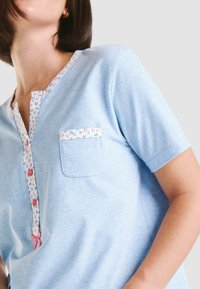 Camisa de algodón azul claro con mangas cortas, cuello con un patrón floral y ribete en el bolsillo, y botones rosas para el cierre.