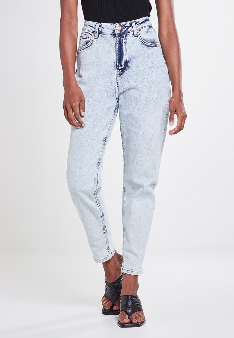 Jean en denim bleu clair taille haute avec une finition légèrement déchirée, dotés de poches avant et d'une fermeture à bouton classique. Texture douce.