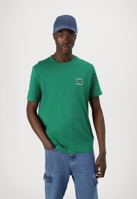 Zöld pamut póló kerek nyakkal, fehér "Tommy Hilfiger" felirattal, kék sapkával és farmer cargo nadrággal párosítva.