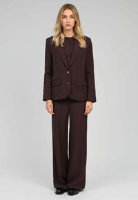 Femme aux cheveux blonds portant un costume marron foncé ajusté avec un pantalon large et des chaussures noires, se tenant devant un fond gris clair uni.